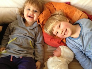 Foto von den zwei Geschwistern, liegend mit ihrem Neugeborenem auf der Couch